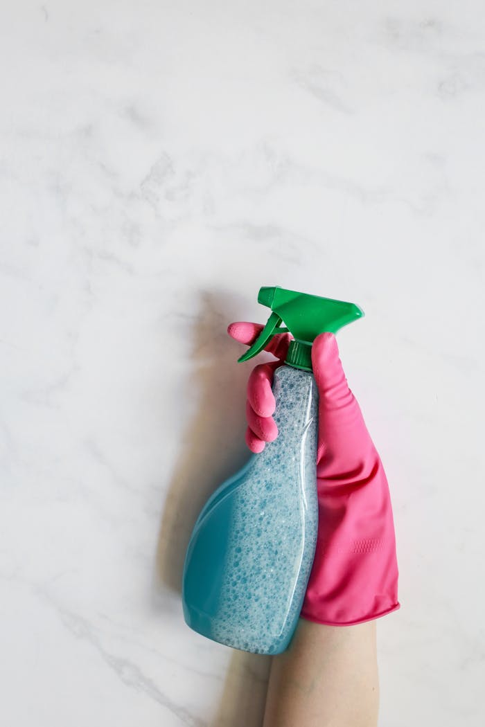 Close-up image of a hand in a pink glove holding a spray bottle against a white background.
