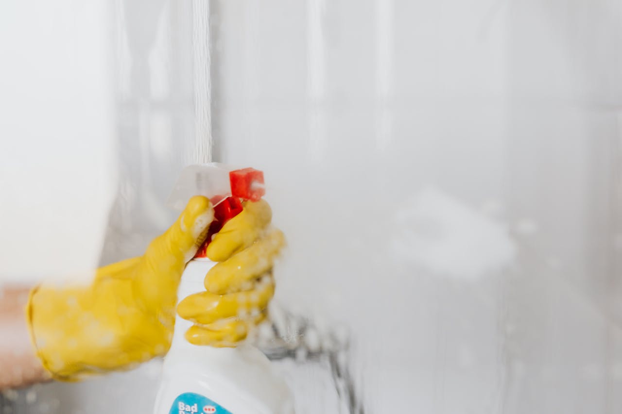 Crop unrecognizable person in yellow latex protective glove spraying detergent from bottle with plastic dispenser at glass of shower cabin